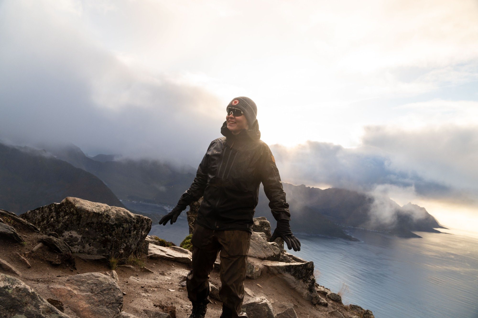 Smiling in rain gear on a rocky summit, with clouds over the fjord behind.